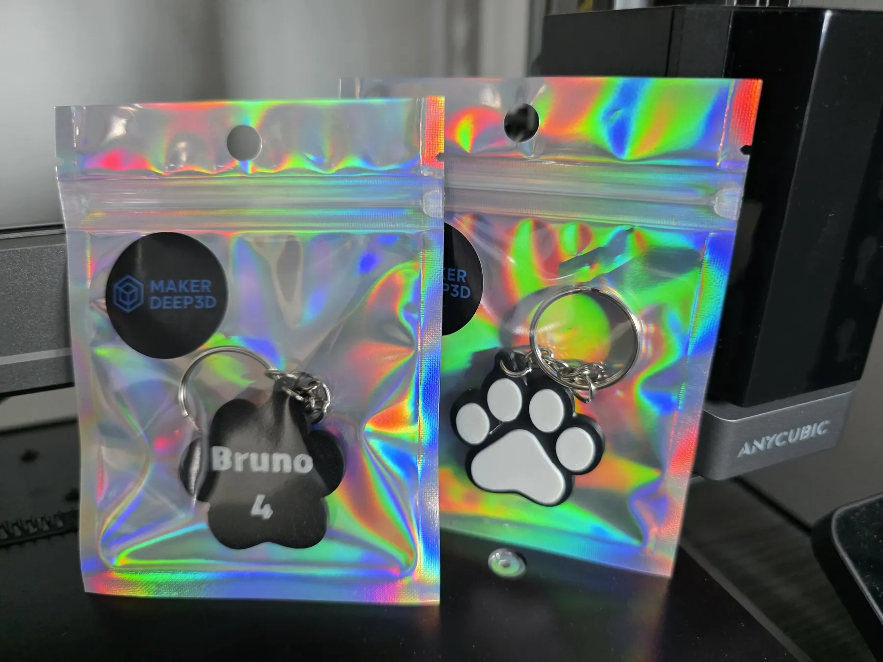 Dos bolsas holográficas con sello de MakerDeep3D: una con una placa negra grabada y otra con una placa en forma de huella